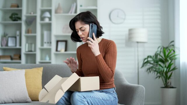 Disappointed young adult woman on couch holding phone, expressing frustration about online shopping issue. Open box in front means delivery problem. Unhappy female receiving parcel with wrong product