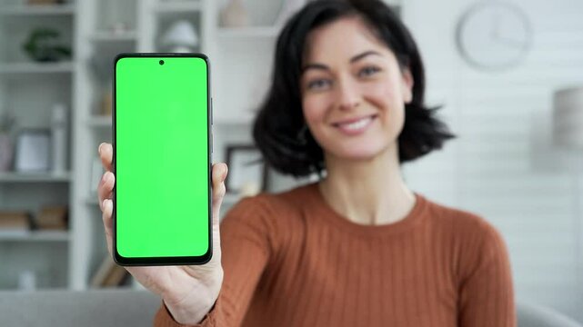 Happy young woman showing mobile phone with green screen sitting on sofa in living room at home. Color key, vertical template layout for advertising. Smiling female looking camera holding smartphone
