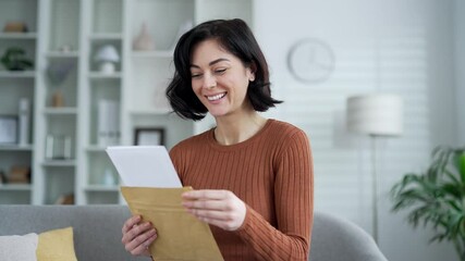 Joyful young woman smiling while holding envelope and reading letter in modern apartment. Happy excited female received great news with happiness and excitement. The lady holds a positive notice