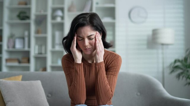 Young adult woman suffering from headache while sitting on sofa in living room at home. Upset female massages her temples with her hands. She is tired, feels pain, needs treatment. Stress and fatigue