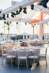 Elegant Waterfront Outdoor Wedding Reception with Draped Canopy, Floral Centerpieces, and White Chairs at a Luxurious Seaside Venue