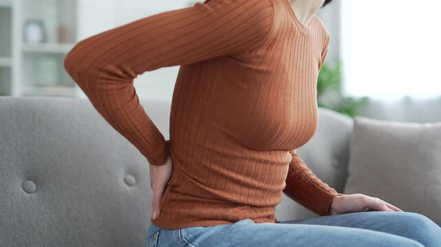 Close up. Female hand holding massaging lower back. Tired young adult woman suffering from back pain while sitting on sofa in living room at home. Upset lady shows discomfort due to poor posture