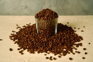 A glass jar filled with roasted coffee beans, with scattered coffee beans around it on a light surface.