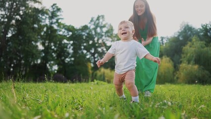 Fototapeta premium Mom and baby taking the first steps. Happy family kid dream concept. A baby is walking on the grass in a park. Mom and baby walking on grass on first steps in the lifestyle park.