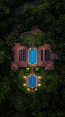 Aerial view of a luxurious property featuring two pools surrounded by lush greenery, illuminated by soft lights at night.