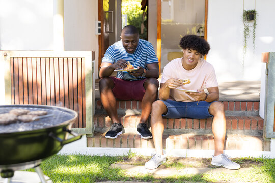 Eating burgers and relaxing, two diverse male friends enjoying outdoor barbecue together