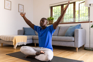 At home, Meditating on yoga mat, african american man raising arms and sitting cross-legged in livin