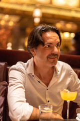 Man drinking cocktail in Bar. A man in a white shirt sitting at a bar or restaurant, smiling slightly with a drink on the table. The background is warmly lit, creating a cozy atmosphere.