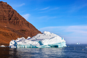 Blue iceberg, red rock mountain and blue sky. A clear day in the cold fjord waters of Scoresby Sund, Greenland. © Rixie