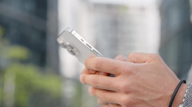 Close camera view of man hands holding phone while standing outdoors. Typing message with fingers. Tapping on touch screen. Chatting online in application or social media. Black bracelet on male arm.