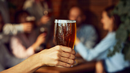 Raised glass of beer in focus, while group of friends in blurred background cheer and celebrate, likely in cozy pub. Emotions joyful and festive. Concept of Oktoberfest, brewery, vacation, Friday mood
