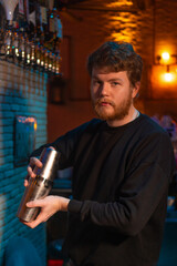 Professional bartender with a beard in a black sweatshirt holds a steel shaker behind the bar and looks at the camera. Bartender in a beautiful and atmospheric atmosphere in the bar. Vertical photo