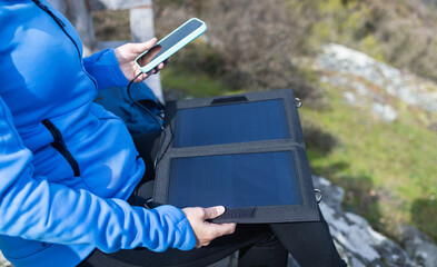 Young woman connecting her smartphone to solar panel charger outdoor after mountain trekking -...