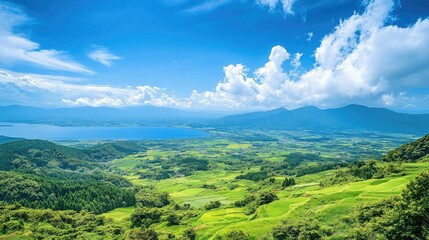 Obraz premium A panoramic view of Kurumayama Plateau with lush green fields, Lake Shirakaba glimmering in the distance, and soft clouds drifting across a blue sky.
