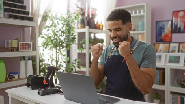 Young hispanic man celebrating a happy win with a smile while using laptop in apron at a home decor store - achievement and joy in every expression