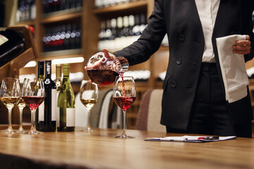 Woman bartender in strict classical clothes pouring red wine from decanter to the glass, close up...