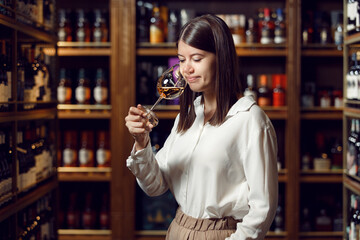 Young professional female sommelier tasting wine standing in cellar on bottles shelf background, close up. Wine expert smelling white wine into glass. 