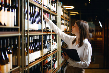 Professional woman sommelier getting wine bottle from shelves, making notes on tablet being at wine cellar.