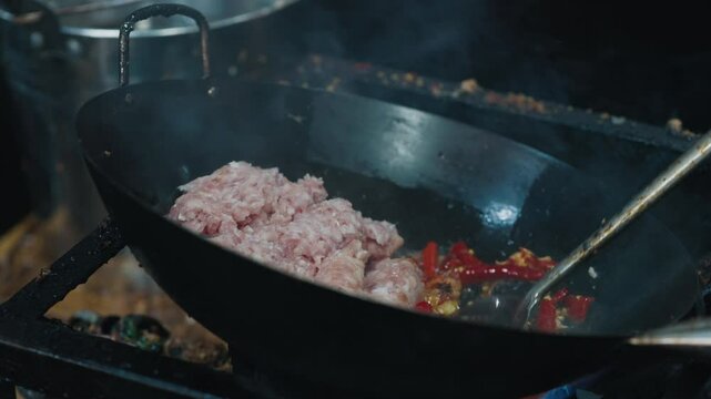 Unrecognizable kitchener holding deep iron pan at angle while frying red chili peppers in fat and juice from grounded pork. Original recipe for main meat dish with bright taste and aroma.