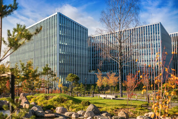 A sleek glass building reflects the sky and clouds, framed by green grass, autumn trees, and a...