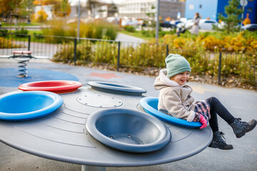 A cute little girl sitting on a round, grey-colored playground spinner with colorful seats