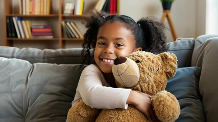 A young girl smiles as she hugs her teddy bear while sitting on a couch in a living room - Powered by Adobe