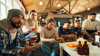 Thrill of game takes over as guys lean in, giving it their all. Perfect blend of tension and laughs as they share drinks and play. Concept of football fans, sport evens, video games and cybersport.