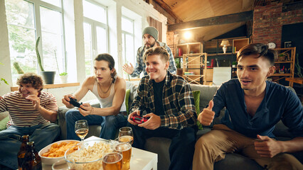 Guys, fully engrossed in game, sit together in cozy space holds controllers in hand, while chips and popcorn sit untouched. All focus on score. Concept of football fans, sport evens, video games.