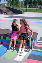 Two preschool-age girls relax and have fun chatting in a skatepark after skateboarding