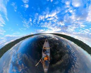 A very wide-angle view looking down on a man paddling a canoe on a large calm lake under a blue sky...