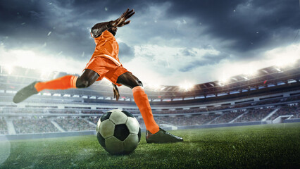 Athletic young African-American man in orange uniform, soccer player in motion on outdoor arena, dynamically playing, showing readiness to score goal. Concept of sport, tournament, competition, game
