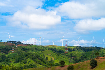 A panoramic view of a wind farm nestled amidst rolling green hills. White wind turbines stand tall against a backdrop of blue sky dotted with fluffy clouds. The scene is both serene and powerful,