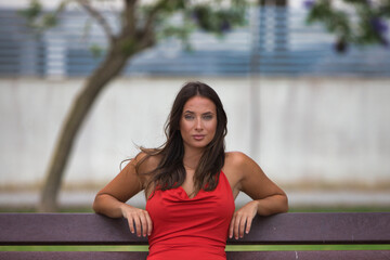 Beautiful young Spanish woman with long hair and green eyes wearing a very elegant red dress sitting on a wooden bench in the park. The woman has her legs crossed and is resting peacefully.