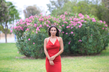 young and beautiful Spanish woman with long hair and green eyes dressed in a very elegant red dress walks like a model on the grass in the park and does different body postures.