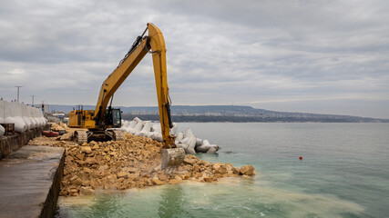 Fototapeta premium Waterfront construction site with excavator. Excavator working on a breakwater construction