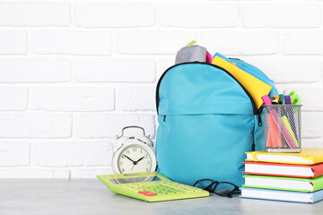Backpack with different school stationery and alarm clock on light grey table near brick wall....