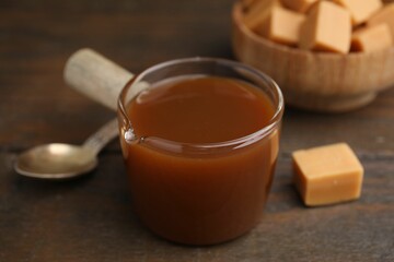 Tasty caramel sauce in glass measuring cup and sweet candies on wooden table, closeup