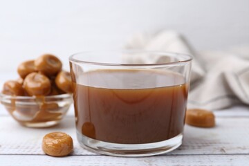 Tasty caramel sauce in bowl and candies on white wooden table, closeup
