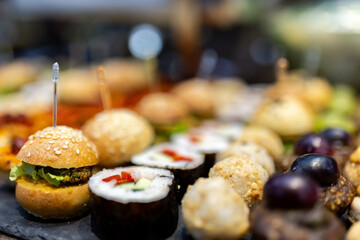 A colorful platter featuring sushi, mini burgers, and assorted delicacies at a vibrant food market in the evening
