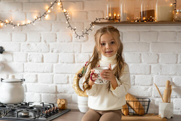 A little girl has fun sitting on kitchen counter and drinking hot tea or chocolate with...
