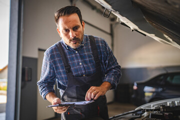 Adult man car mechanic inspect car engine, hold clipboard in workshop