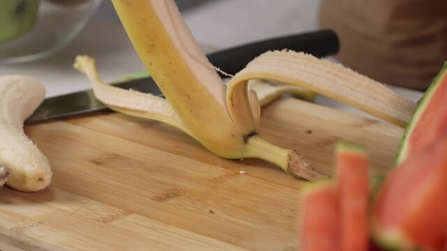 Hands of young woman european peels banana on cutting board for fruit salad, right side view close up. Healthy lifestyle concept, raw food diet, cooking at home, refreshing