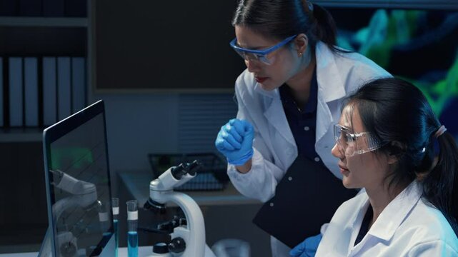 Two female scientists wearing lab coats and safety glasses are analyzing data on a computer screen in a laboratory setting