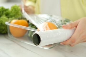 Woman putting plastic food wrap over glass container with fresh vegetables at white marble table,...