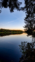 Nature reserve Pláničský pond in Černá v Pošumaví in the evening. The calm water mirrors the surrounding trees and sky. A bench and a small boat are visible by the shore. 