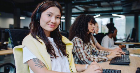 Beautiful Asian woman cheerfully smiling while getting distracted by camera from work. Working in modern office. People wearing headsets for remote consultation with customers. Using computers.