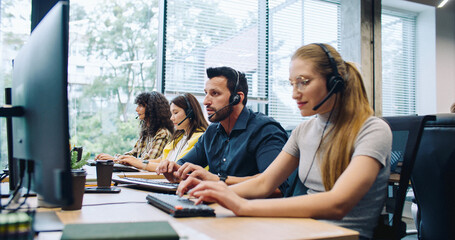 Hardworking dispatchers or consultants actively talking with clients on headsets. Attractive leader walking in background and controlling work process. People using computers at work. Support center.
