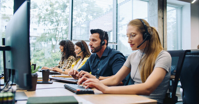 Hardworking dispatchers or consultants actively talking with clients on headsets. Attractive leader walking in background and controlling work process. People using computers at work. Support center.