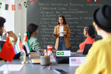 Young female teacher standing by blackboard and explaining topic to students during lesson in...