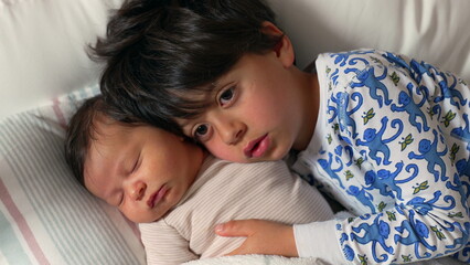 Big brother rests his head against his sleeping newborn sibling, both lying together on a bed, wrapped in soft blankets, sharing a peaceful and loving moment